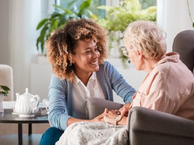 A smiling caregiver holds the hands of an elderly woman.