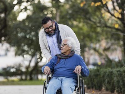 A senior Hispanic man in his 80s sitting in a wheelchair, taking a walk in the park with his adult son, a mid adult man in his 30s.