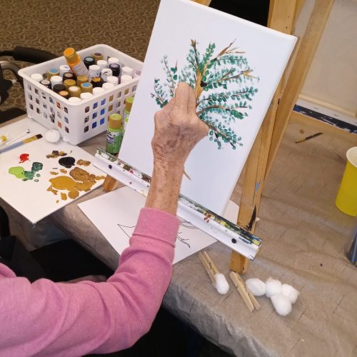 A senior woman participates in art therapy, painting a beautiful and relaxing picture of a tree.